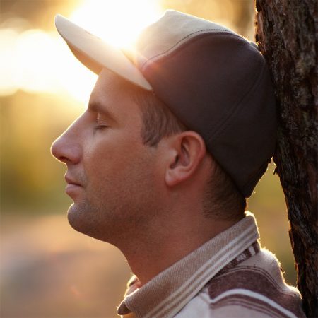 Young man meditating outdoors, in profile, leaning against tree